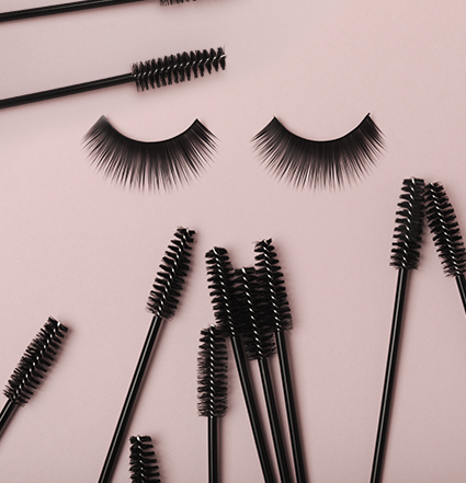 Two eyelashes at the center, surrounded by mascara wands on a pinkish background
